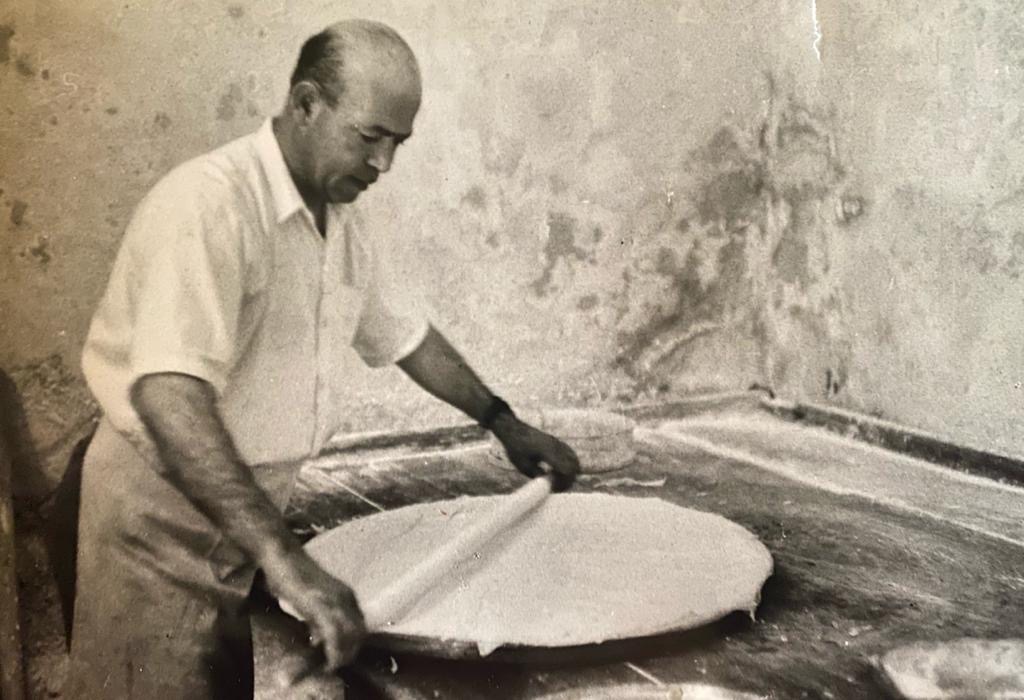 Black-and-white photograph of a man rolling a large round of paper-thin mutabbaq dough on a marble slab
