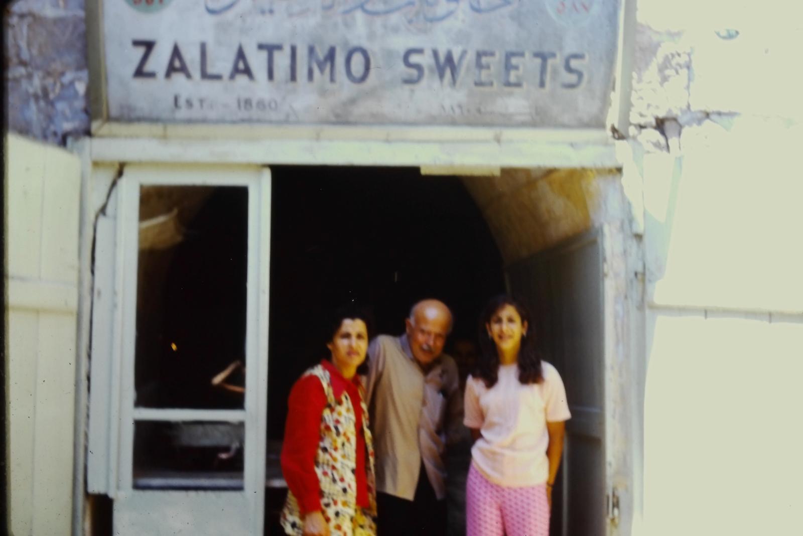 Color photograph of three people standing beneath the Zalatimo Sweets, Est. 1860 sign at the shop entrance